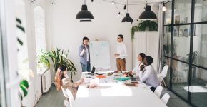 High angle of multiethnic team gathering in modern conference room with glass wall while male coworkers standing near flip chart presenting financial statistics