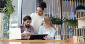 Young people man and women coworking together at creative stylish office sitting at the table boss checking project ideas while women looking at him holding paper holder smiling cheerful.